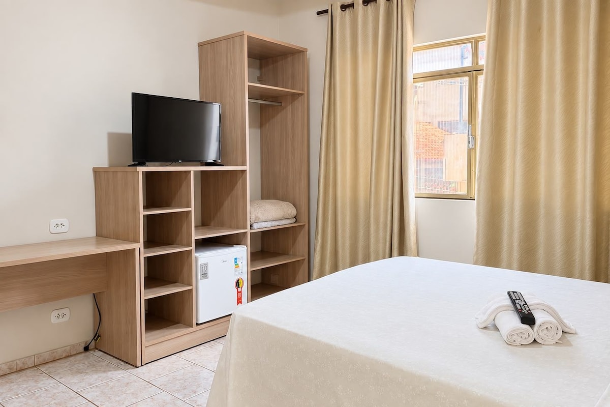 Habitación de hotel con cama, televisión sobre una estantería de madera, un refrigerador pequeño y una ventana con cortinas.