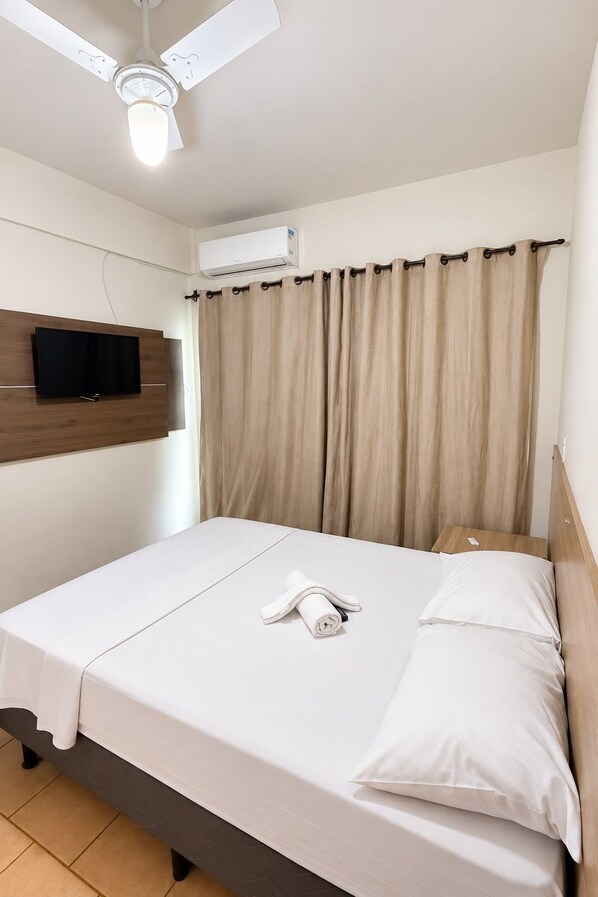 A hotel room with a bed, a flat-screen TV mounted on the wall, an air conditioning unit, and a ceiling fan.