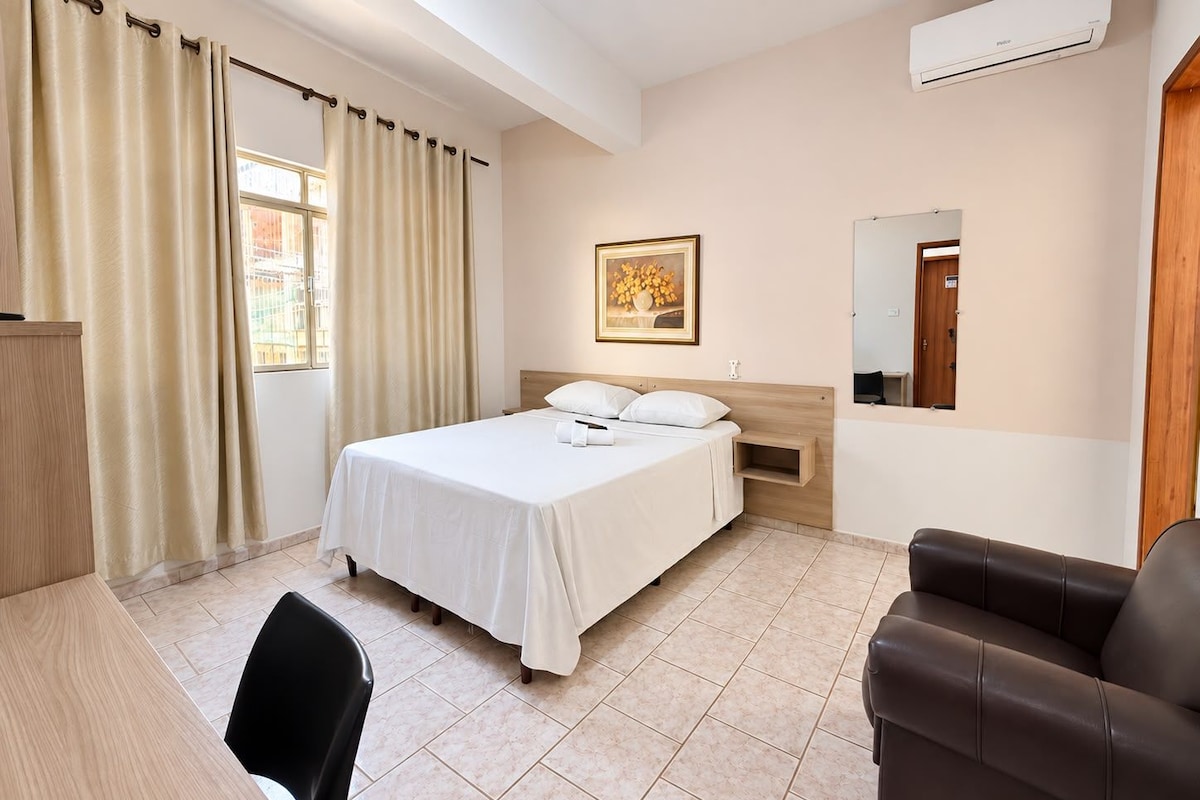 Une chambre d’hôtel comprenant un lit, un bureau, une chaise, un canapé en cuir marron, une fenêtre avec des rideaux, un tableau au mur et un appareil de climatisation.