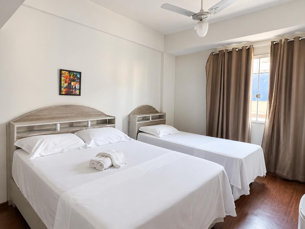 Ein Hotelzimmer mit zwei Betten, einem Deckenventilator, einem Fenster mit Vorhängen und einem Gemälde an der Wand.