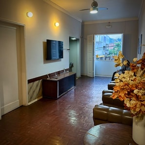 A spacious living area with a flat-screen TV mounted on the wall, a wooden floor, a seating area with a sofa and a coffee table, and a view of the outdoors through a door.