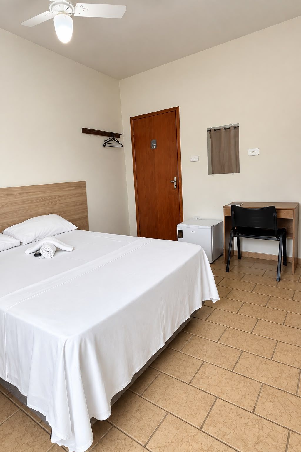 A hotel room with a bed, a desk, a chair, a ceiling fan, a door, and a towel on the bed.