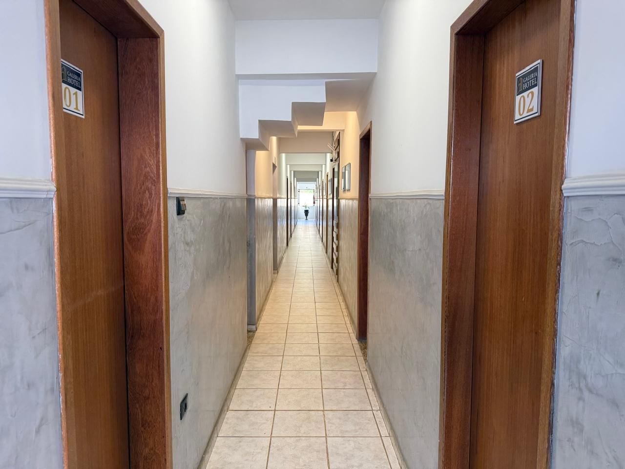 Corredor de hotel com portas numeradas 01 e 02, dando acesso aos quartos.