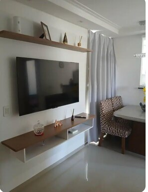 Ein modernes Wohnzimmer mit einem an der Wand befestigten Flachbildfernseher, einer Holzunterhaltungseinheit darunter, einem gemusterten Sessel und einem Essbereich mit einem weißen Tisch.