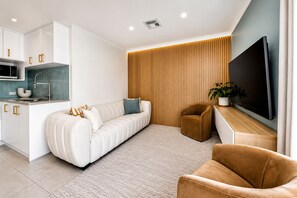 A modern living room with a sofa, armchair, TV, and a kitchen area with cabinets and a sink. - The Duchess of Duke | Spa, Sauna & Beach Walk (Scarborough)