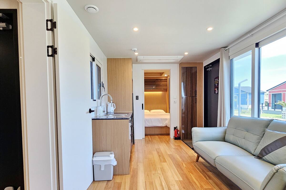 Una habitación de hotel compacta con kitchenette, una cama, un sofá y un ventanal grande.
