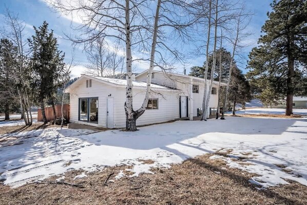A single-story white house with a front door, windows, and a garage door, surrounded by trees and snow. - Woodland Park Home with Direct Views of Pikes Peak! (Woodland Park)