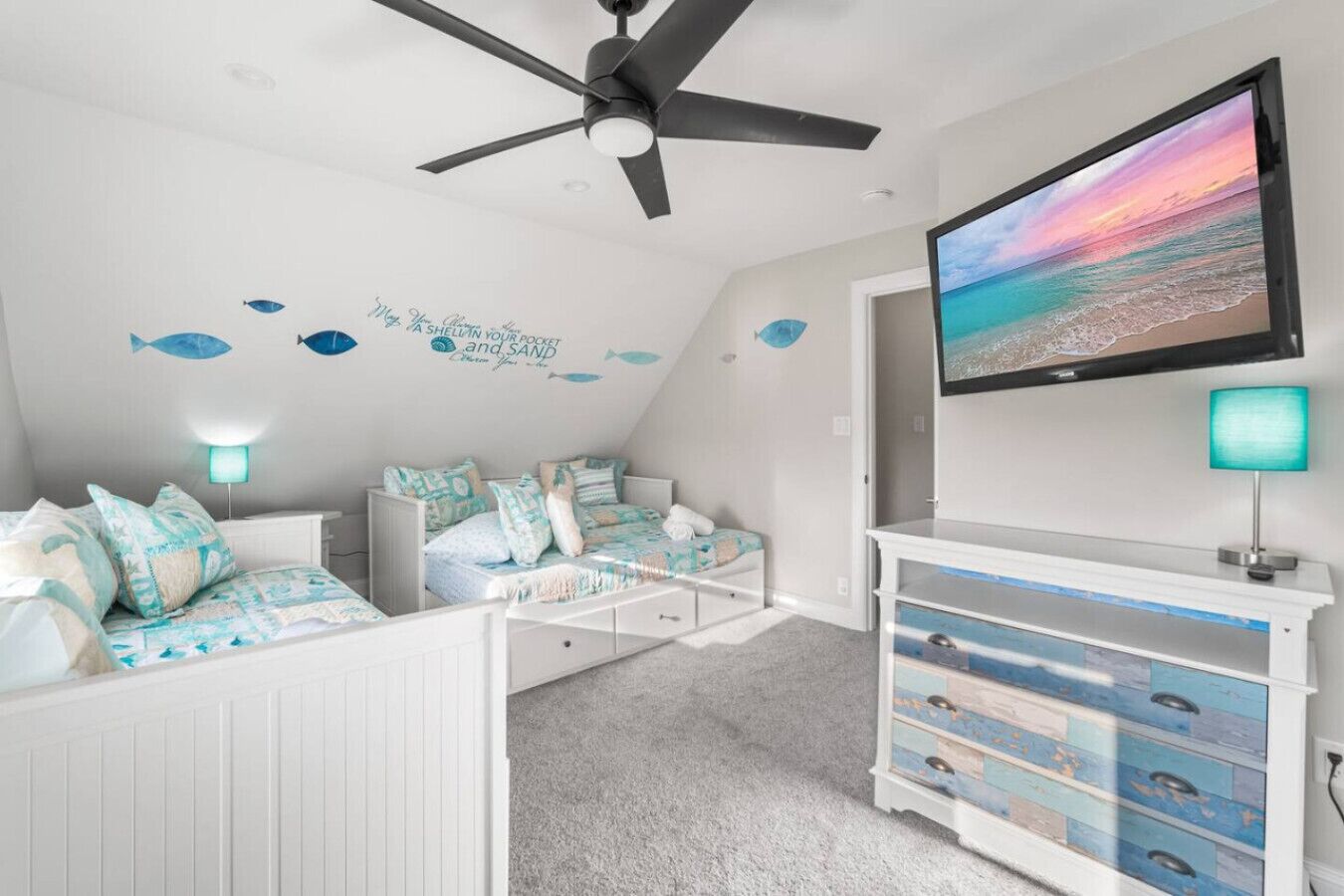 Une chambre dotée d’un ventilateur de plafond, d’un lit recouvert d’une couvre-lit bleue et blanche, d’une commode avec des tiroirs et d’un téléviseur fixé au mur qui diffuse une scène de plage.