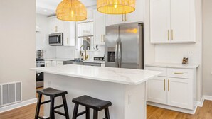 A modern kitchen with white cabinetry, a stainless steel refrigerator, a coffee bar, and two black bar stools. - Modern 8BR Ventnor Compound | Patio | BBQ | Games (Ventnor City)