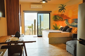 Ein Zimmer mit Bett, einer Couch, einem Esstisch und Blick auf Kakteen und einen Sonnenuntergang.