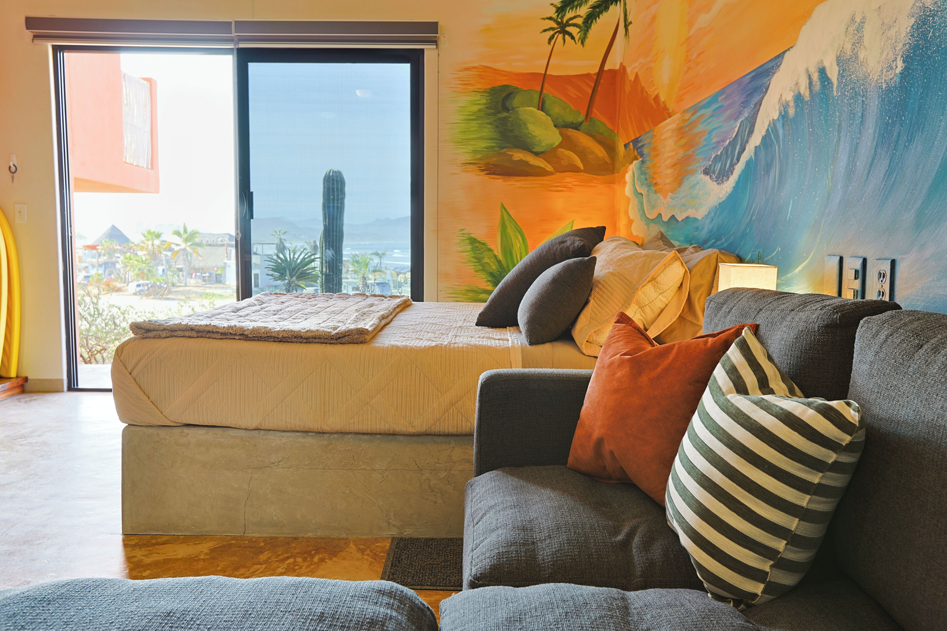 A room with a bed, a sofa, a view through a sliding door, and a mural on the wall.