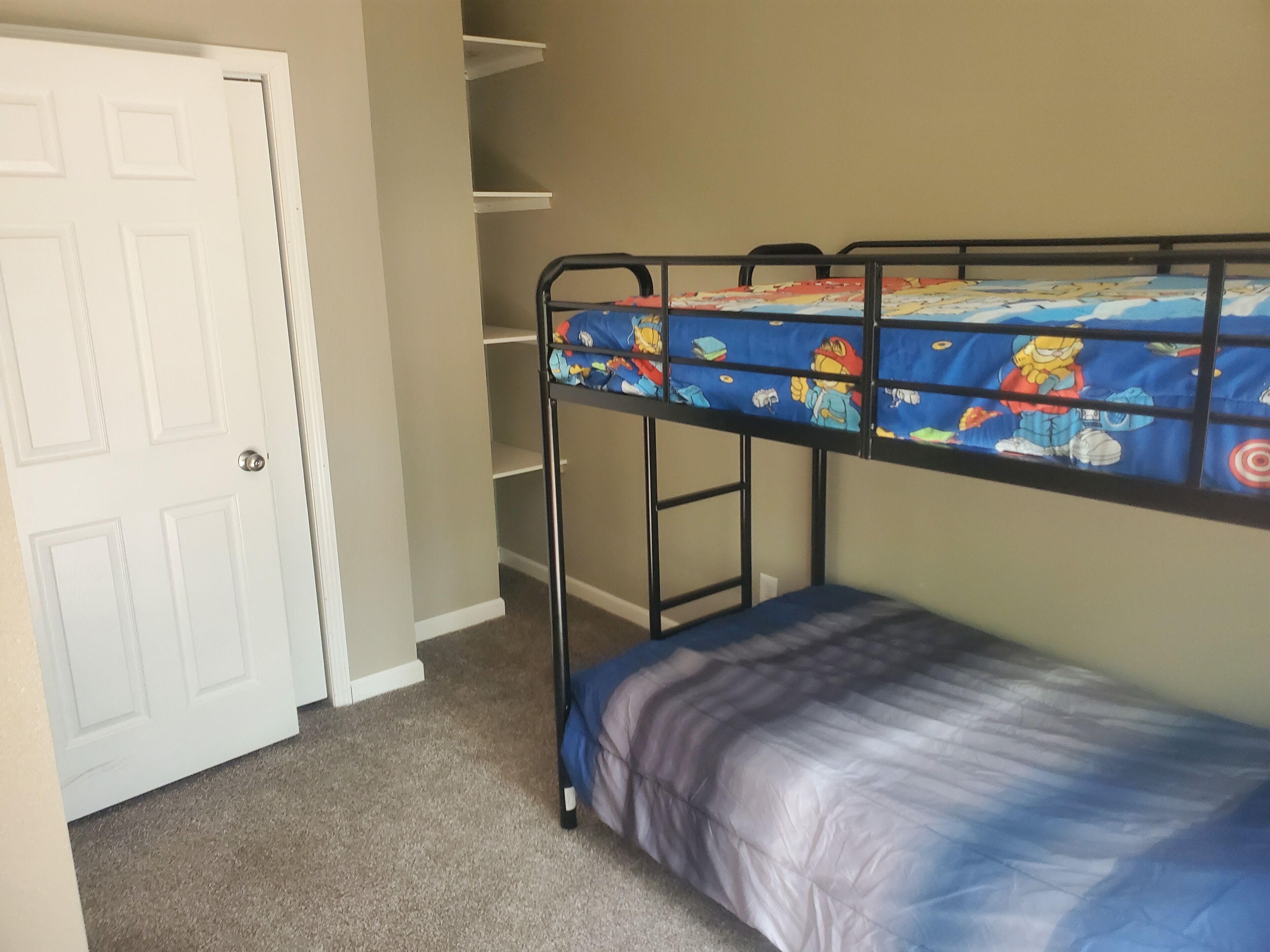 Uma cama de beliche com colchão azul com personagens de desenho animado, uma porta branca e piso coberto de carpete.