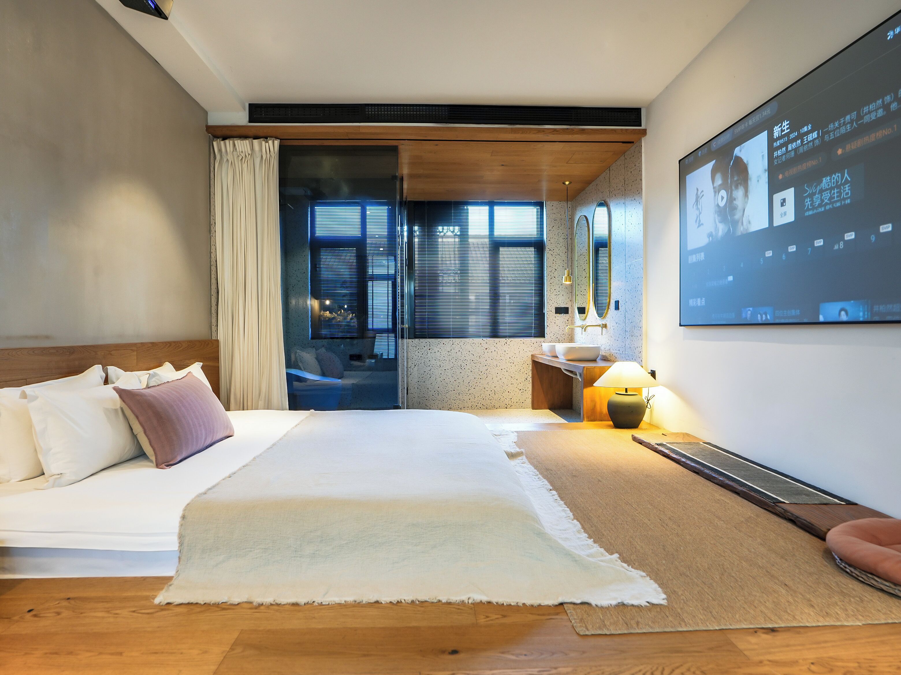 A modern hotel room with a bed, a desk, a large screen, and a view through a glass door.
