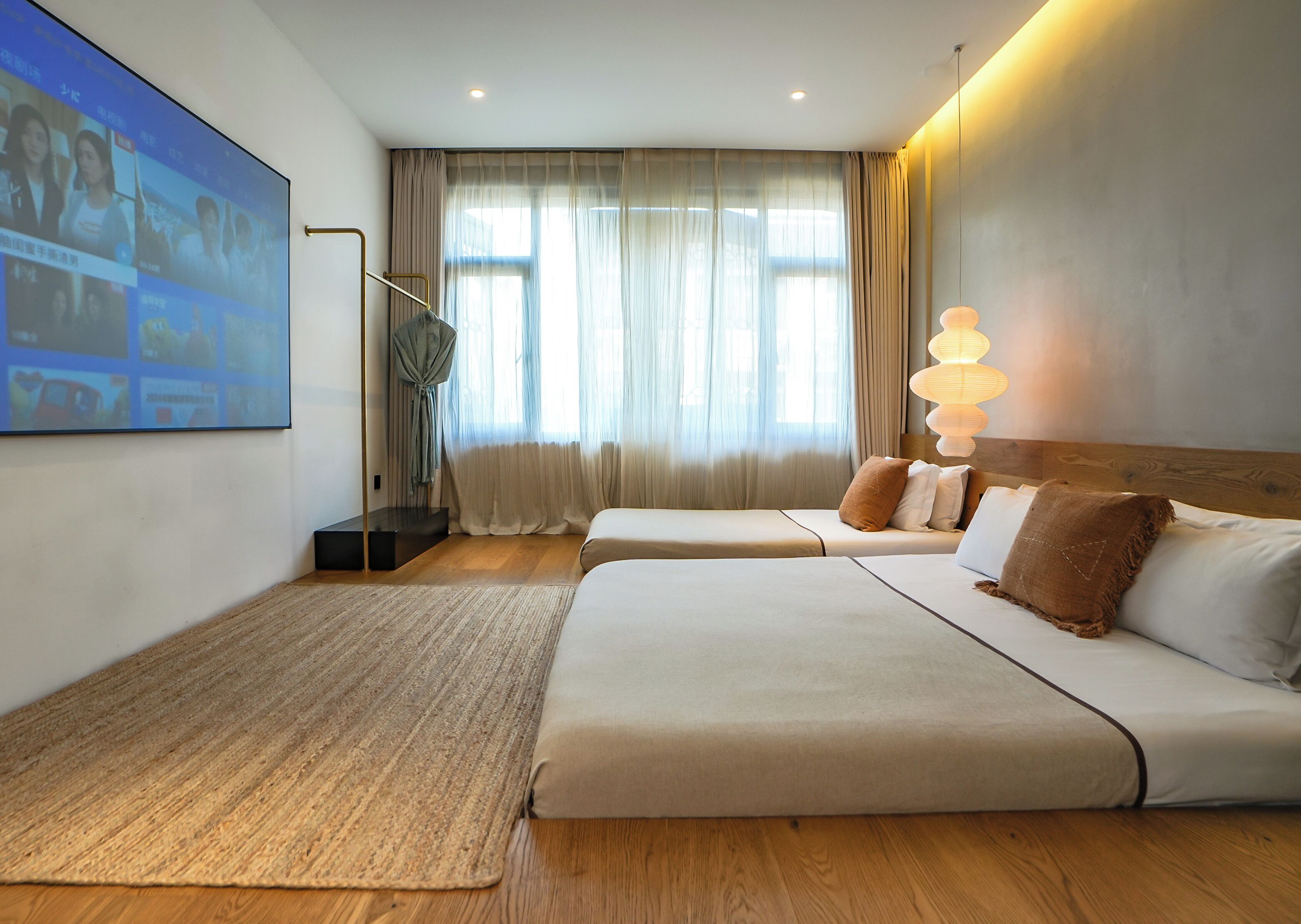 Habitación de hotel con dos camas, una pantalla grande mostrando imágenes, una escalera y vistas al exterior a través de cortinas translúcidas.