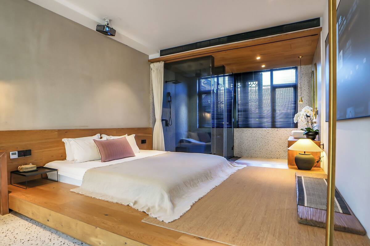 Ett modernt hotellrum med en stor säng, ett nattduksbord, ett skrivbord, en stol, en glasvägg, ett fönster med persienner och en väggmonterad projektor.