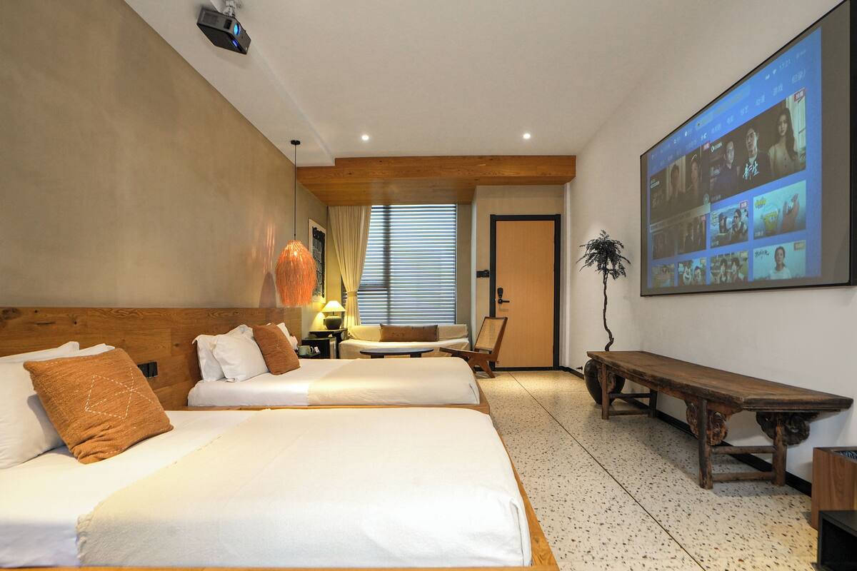 Ett hotellrum med två sängar, ett skrivbord, en stol, en lampa, en växt och en stor skärm som visar ett videokonferenssamtal.