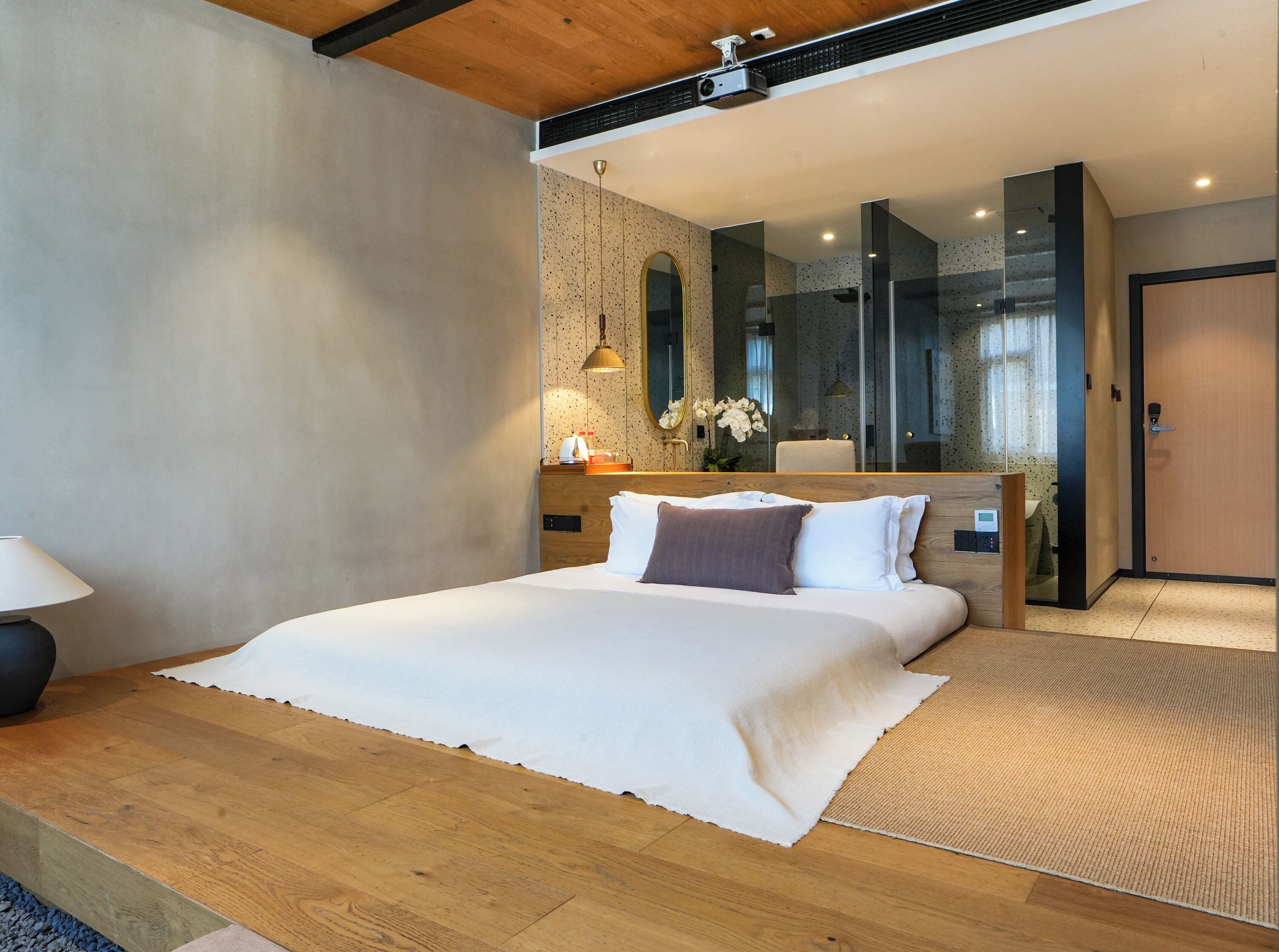 A modern bedroom with a large bed, a wooden headboard, a bedside table with a lamp, and a mirror.