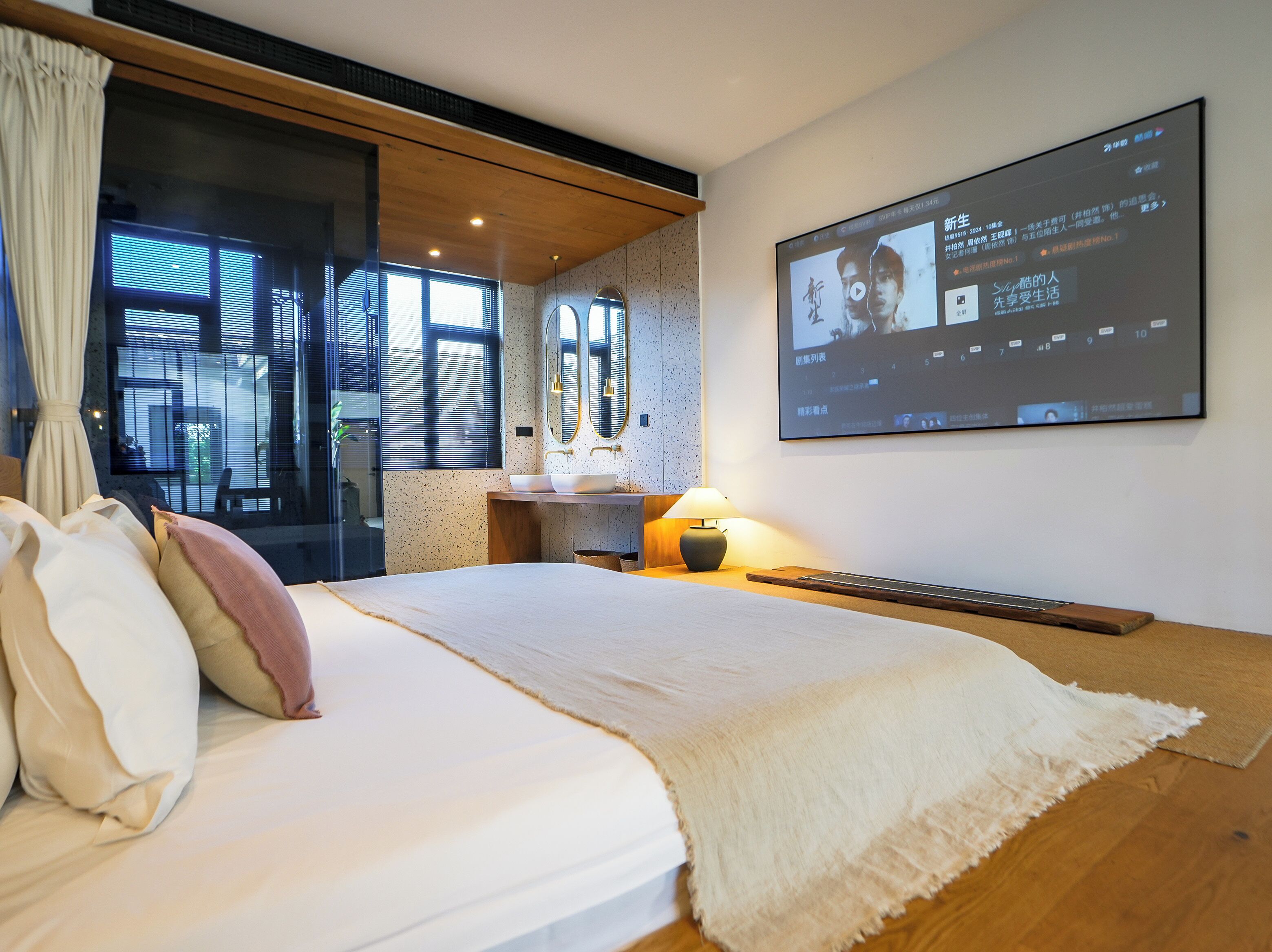 A modern bedroom with a bed, a bedside table with a lamp, a large screen displaying a video, and a window with blinds.