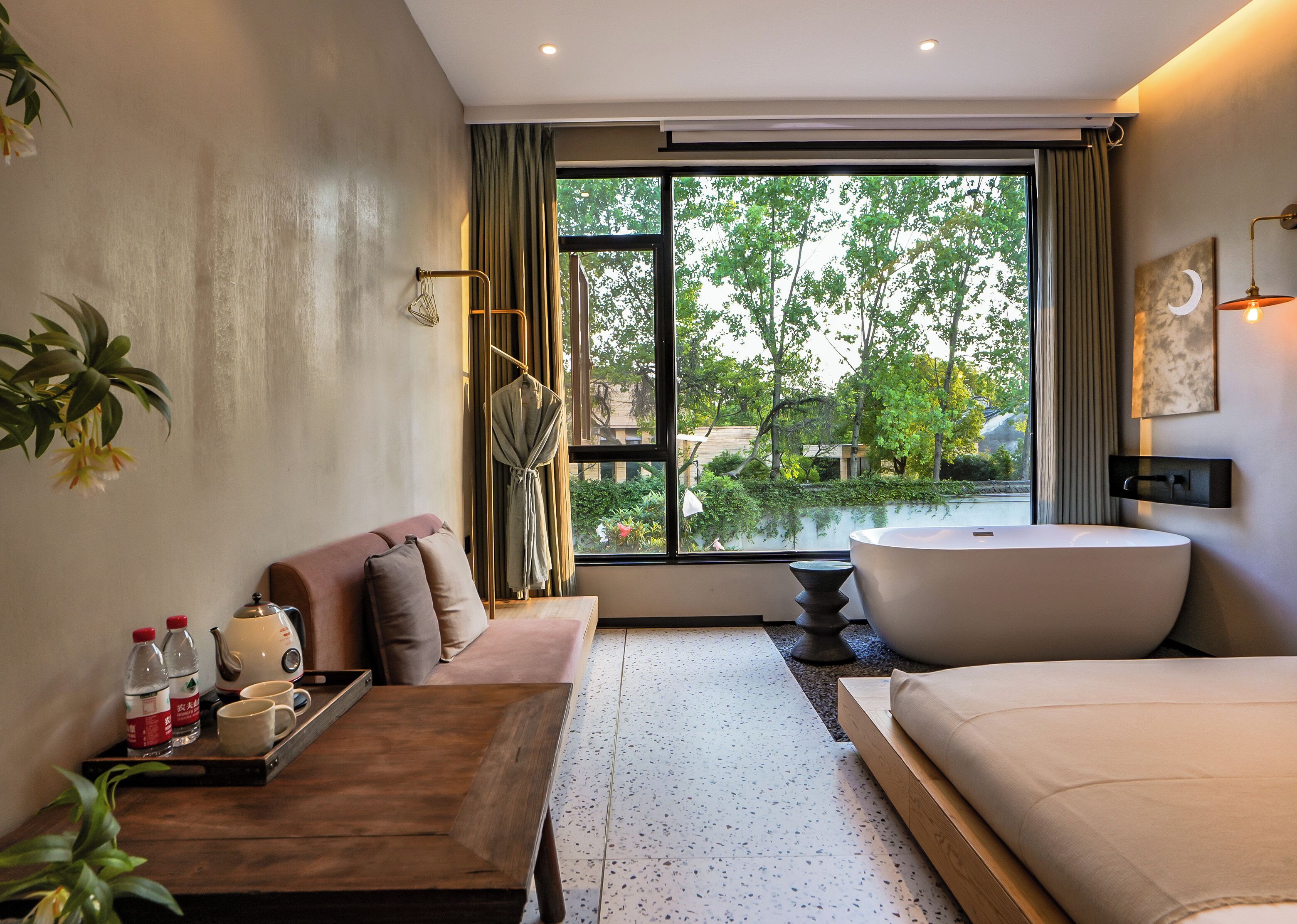 A modern hotel room with a large window, a bathtub, a sofa, a wooden table with bottles and a tray, and a potted plant.
