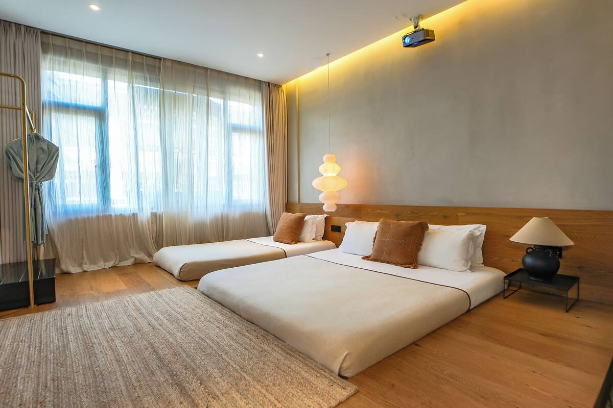 Ett hotellrum med två sängar, ett stort fönster med tunna gardiner, ett trägolv, en golvlampa och en projektor som är monterad på väggen.