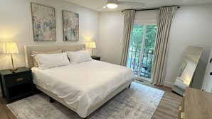 Une chambre moderne avec un grand lit, deux tables de chevet avec des lampes, un ventilateur de plafond, une grande fenêtre avec des rideaux et une vue sur la verdure à l’extérieur.