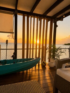 Eine Holzterrasse mit Blick auf den Sonnenuntergang über dem Meer, ein Boot und eine Lounge-Ecke mit einem Stuhl und einer Pflanze.