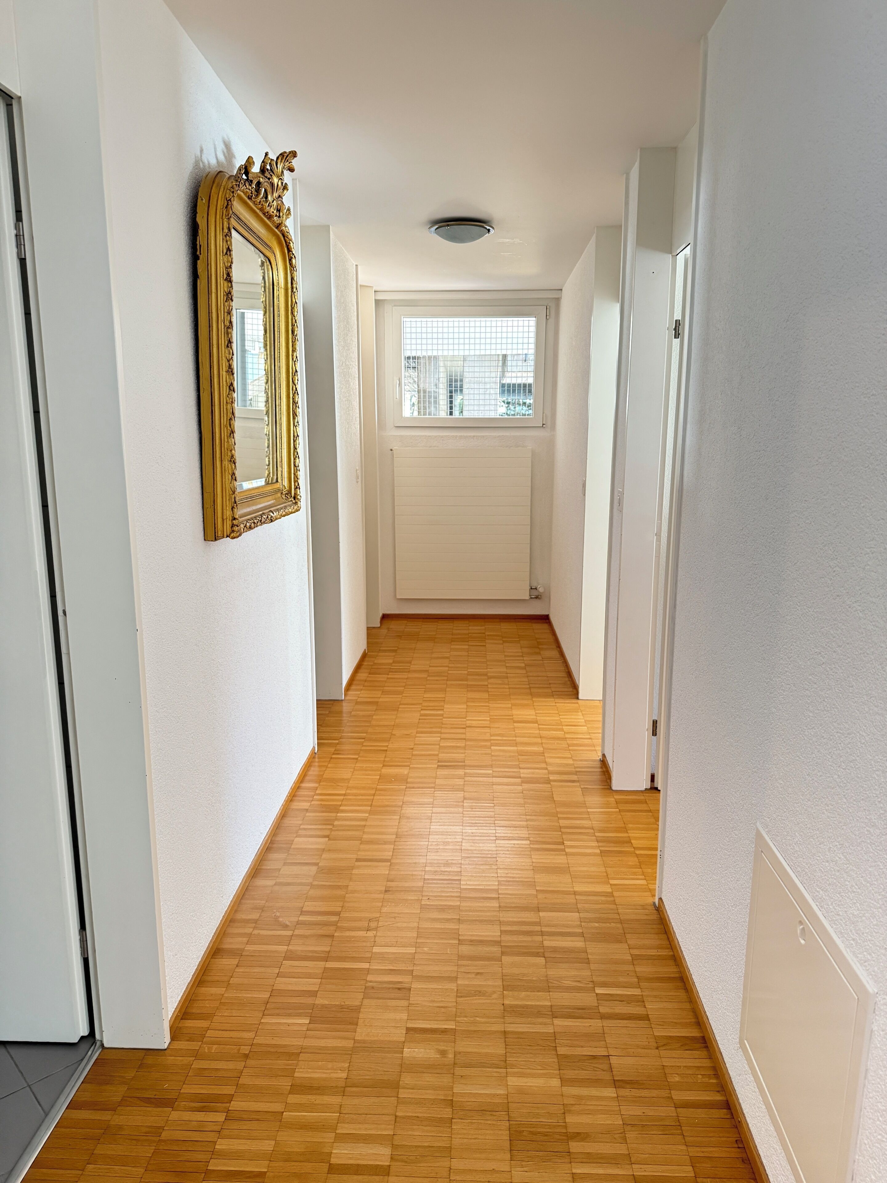 Käytävä, jossa on puulattia, valkoiset seinät, kultakehyksinen peili ja ikkuna, jossa on sälekaihtimet.