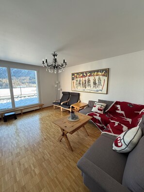 A living room with a wooden floor, a sofa, a coffee table, a bench, and a chandelier. - Terrific 2-Bedroom in St. Moritz Dorf with Stunning Lake View (Sankt Moritz)