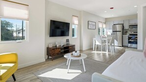 A modern living room with a flat-screen TV, a dining area with white chairs, a kitchen with stainless steel appliances, and a large window. - 2BR Coastal Suite Walk to McGee Beach (Corpus Christi)