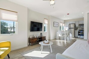 Ein modernes Wohnzimmer mit Flachbildfernseher, einem Essbereich mit weißen Stühlen, einer Küche mit Edelstahlgeräten und einem großen Fenster.