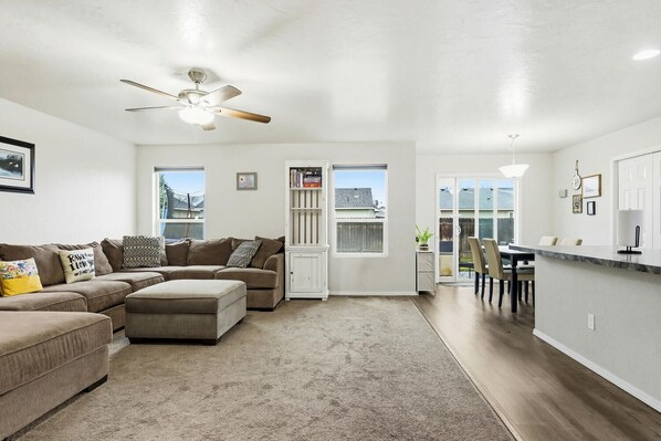 A modern living room with a sectional sofa, a ceiling fan, and a dining area with a kitchen counter.
