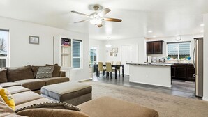 A spacious living room with a sofa, ottoman, and a dining area with chairs. A kitchen with dark cabinets and a refrigerator is visible in the background. - 5 BR Family Fun, Sleep 12, Climbing Wall & Games (Meridian)