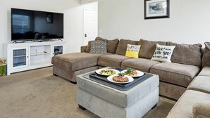 A living room with a sectional sofa, a flat-screen TV, a white entertainment unit, and a grey ottoman with plates of food. - 5 BR Family Fun, Sleep 12, Climbing Wall & Games (Meridian)