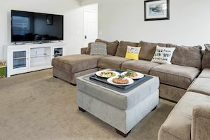 A living room with a sectional sofa, a flat-screen TV, a white entertainment unit, and a grey ottoman with plates of food. - 5 BR Family Fun, Sleep 12, Climbing Wall & Games (Meridian)