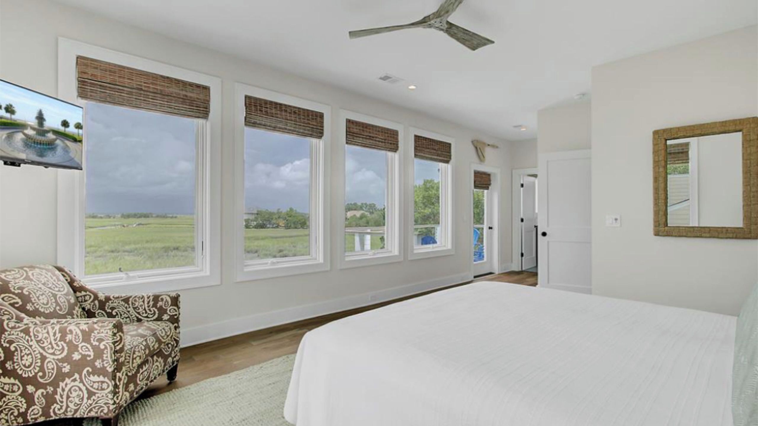 A bedroom with a large bed, a ceiling fan, and a view of a field through large windows.