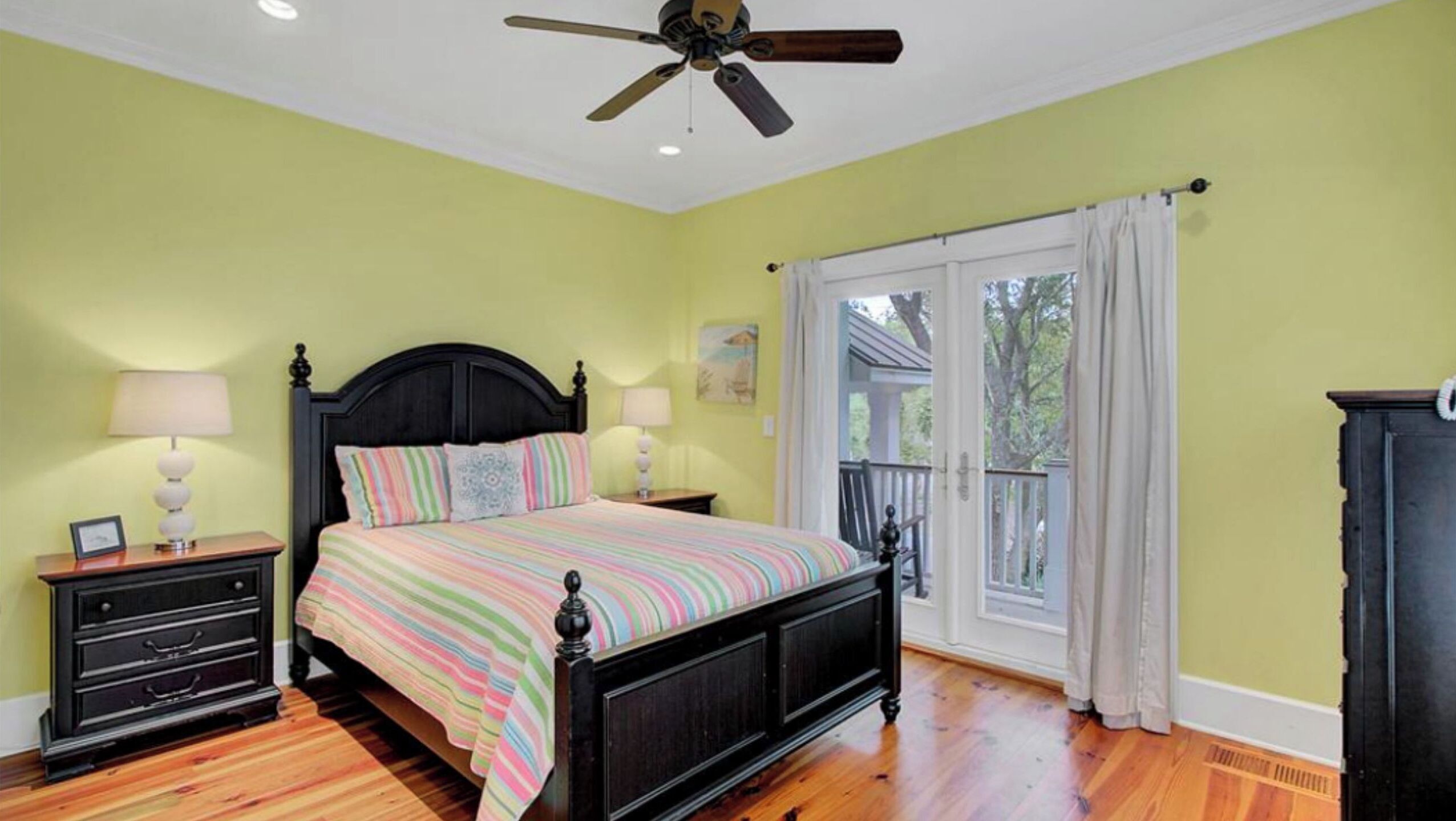A bedroom with a bed, nightstands, a ceiling fan, a window with curtains, and a view of the outdoors.