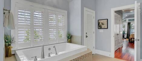 Une salle de bain avec une grande baignoire, une douche et des toilettes. Il y a des fenêtres avec des stores, un porte-serviettes et un tableau encadré au mur.