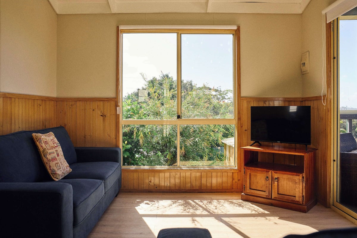 Una habitación con un ventanal grande, un mueble de madera con televisor y un sofá azul oscuro.
