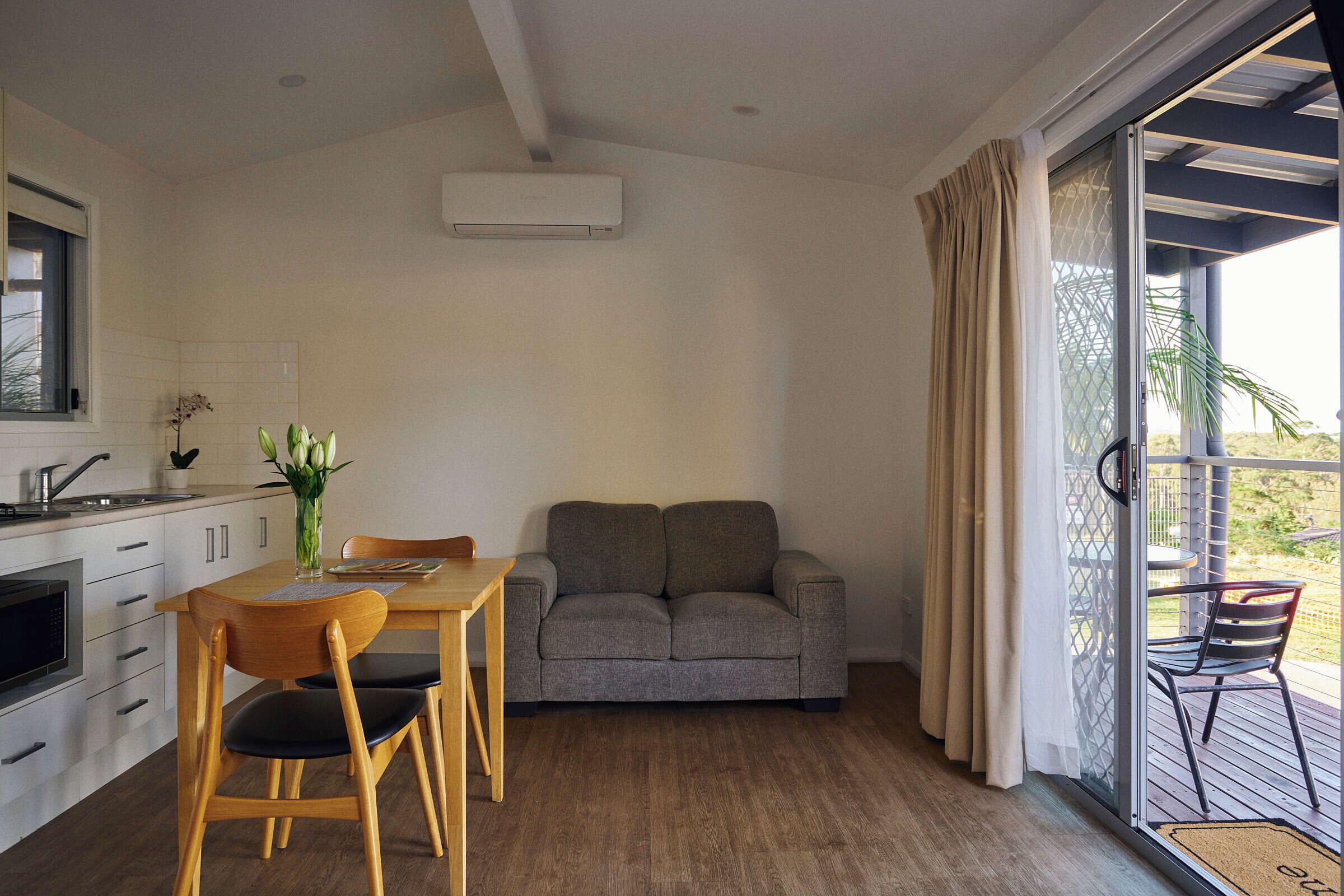 A compact living space with a kitchenette, a grey sofa, wooden dining table, and a sliding glass door leading to an outdoor area.