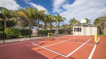 Una cancha de tenis con red, rodeada por una cerca, palmeras y un edificio al fondo.