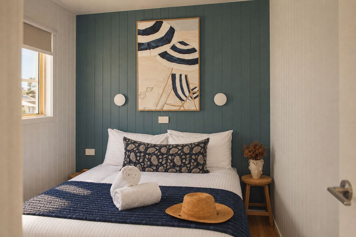 A neatly made bed with a blue and white patterned pillow, a rolled white towel, and a straw hat on the bed.