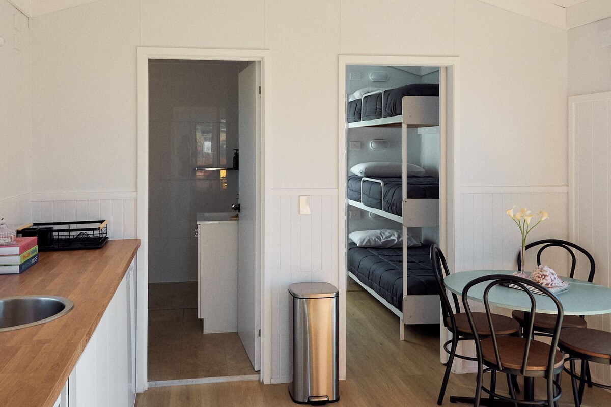 A compact room with bunk beds, a small kitchenette, a dining area with chairs, and a trash bin.