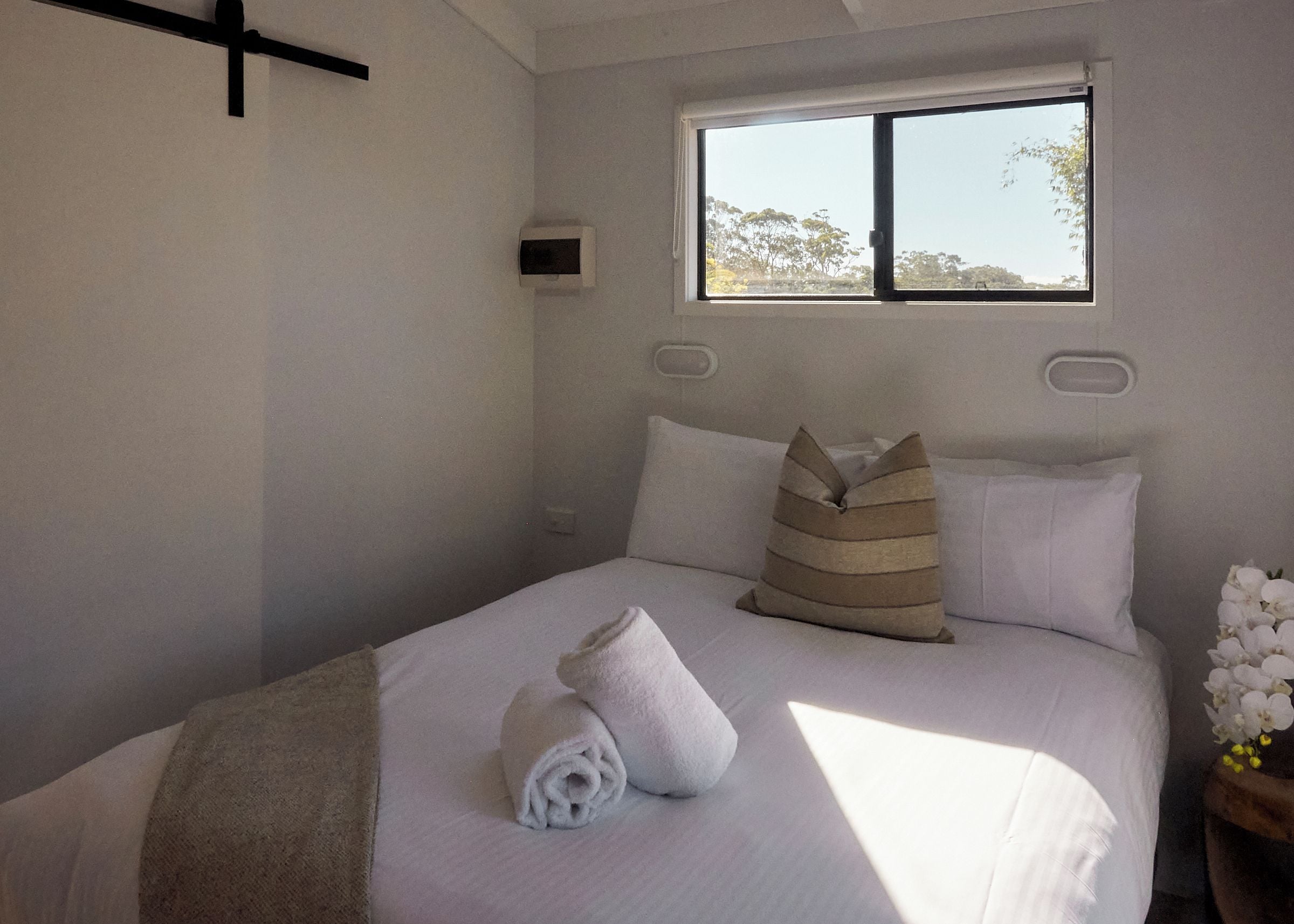 Una cama bien hecha con almohadas, una toalla enrollada y una ventana con vistas a árboles.