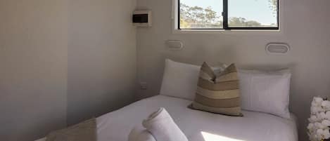 Una cama bien hecha con almohadas, una toalla enrollada y una ventana con vistas a árboles.