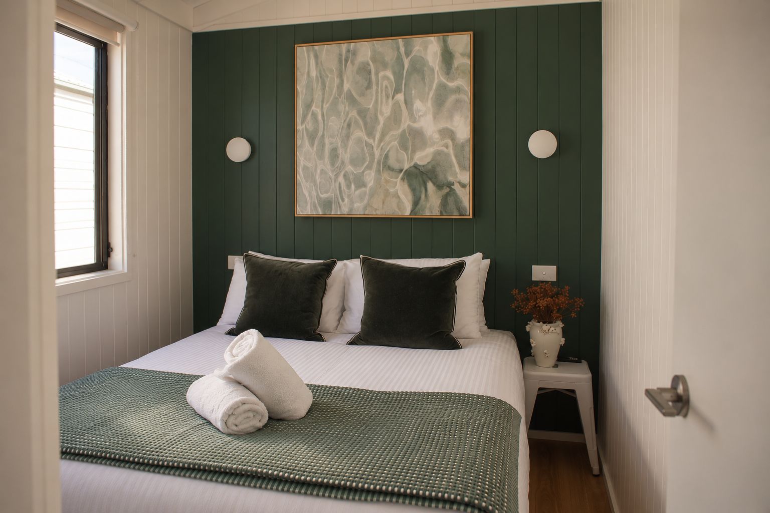 A bedroom with a bed, pillows, a bedside table with a vase, and a painting on the wall.