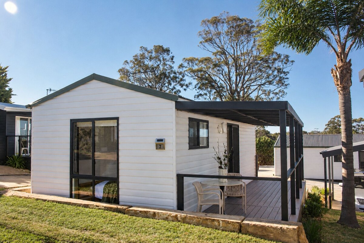 A single-story mobile home with a covered patio area, a small outdoor seating arrangement, and a visible mailbox.