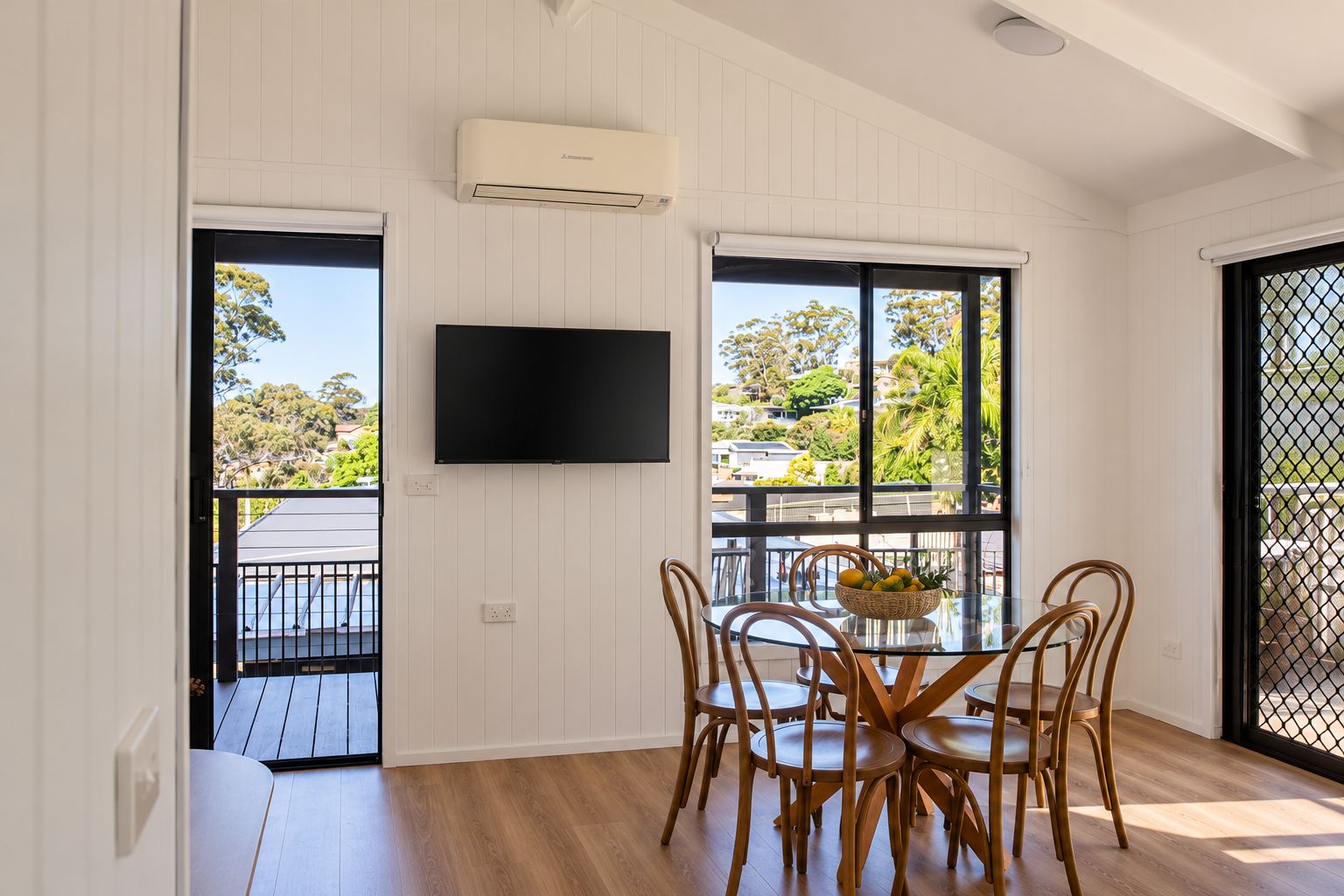 Una habitación con una mesa de comedor, sillas, un televisor y ventanas amplias con vistas a una zona residencial.