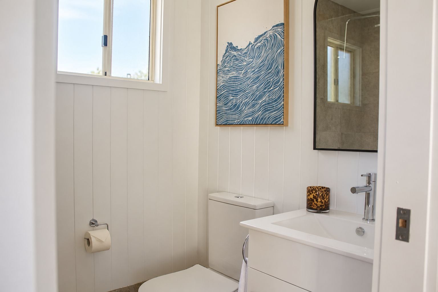 A bathroom with a toilet, sink, and mirror. There is a window and a wall-mounted artwork.