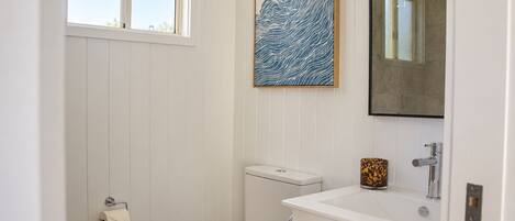 A bathroom with a toilet, sink, and mirror. There is a window and a wall-mounted artwork.