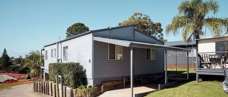 A single-story mobile home with a covered patio, a small front yard, and a neighboring building.